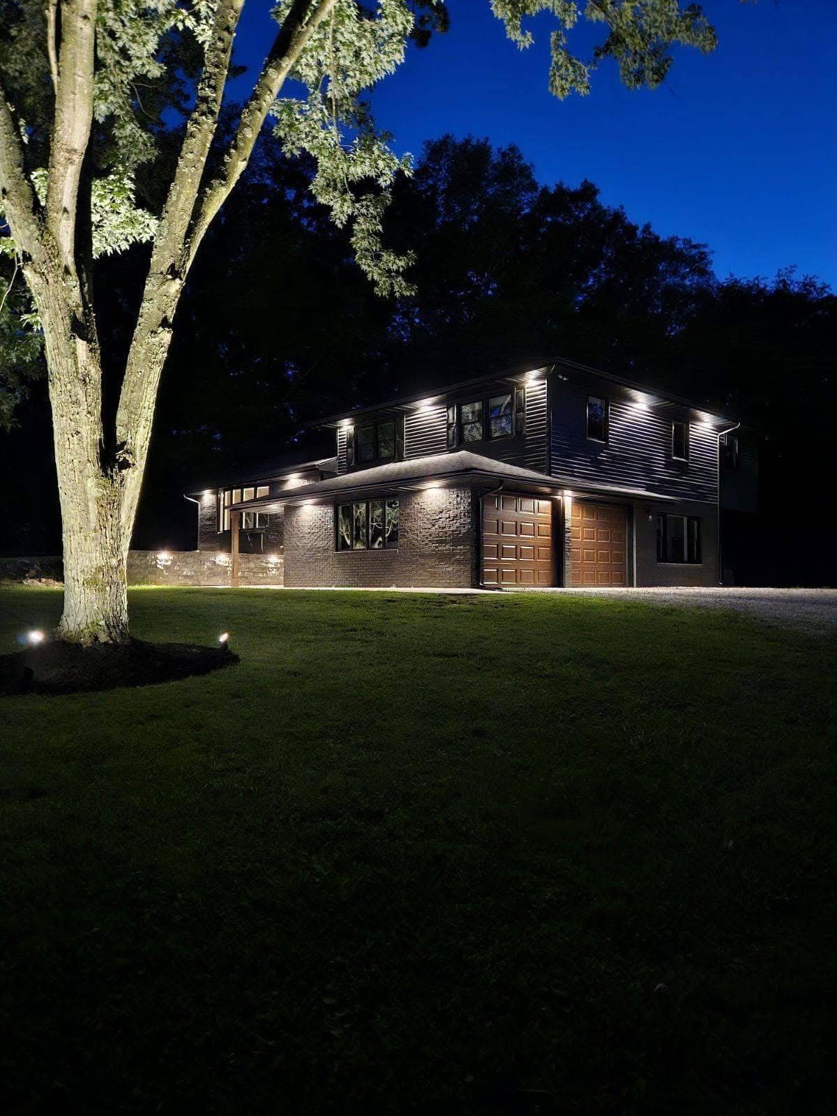 House illuminated at night with spotlights on tree, facade, and ground, surrounded by dark greenery and driveway.