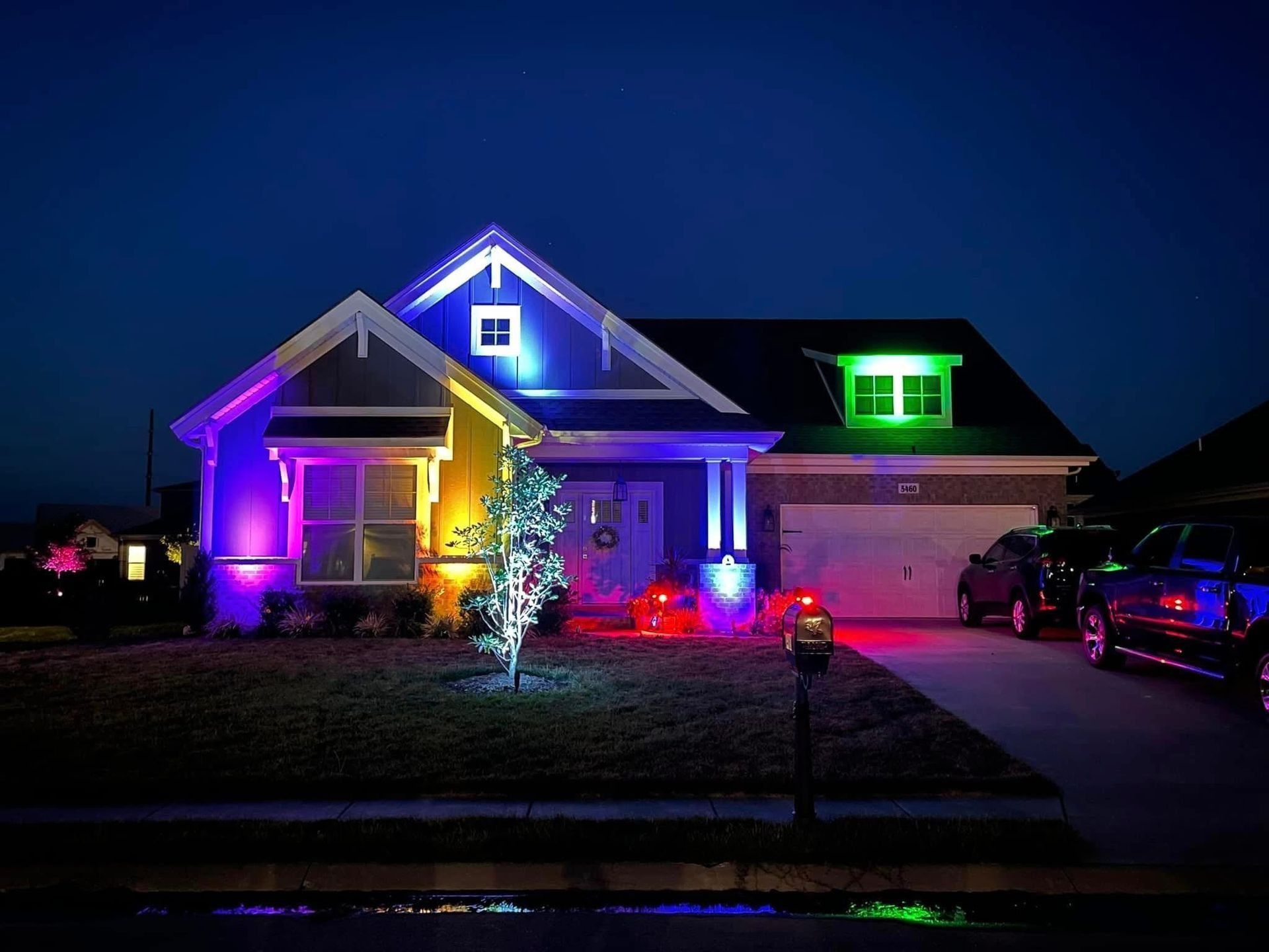 House illuminated at night with colorful lights: purple, green, blue, and red. Driveway with cars.