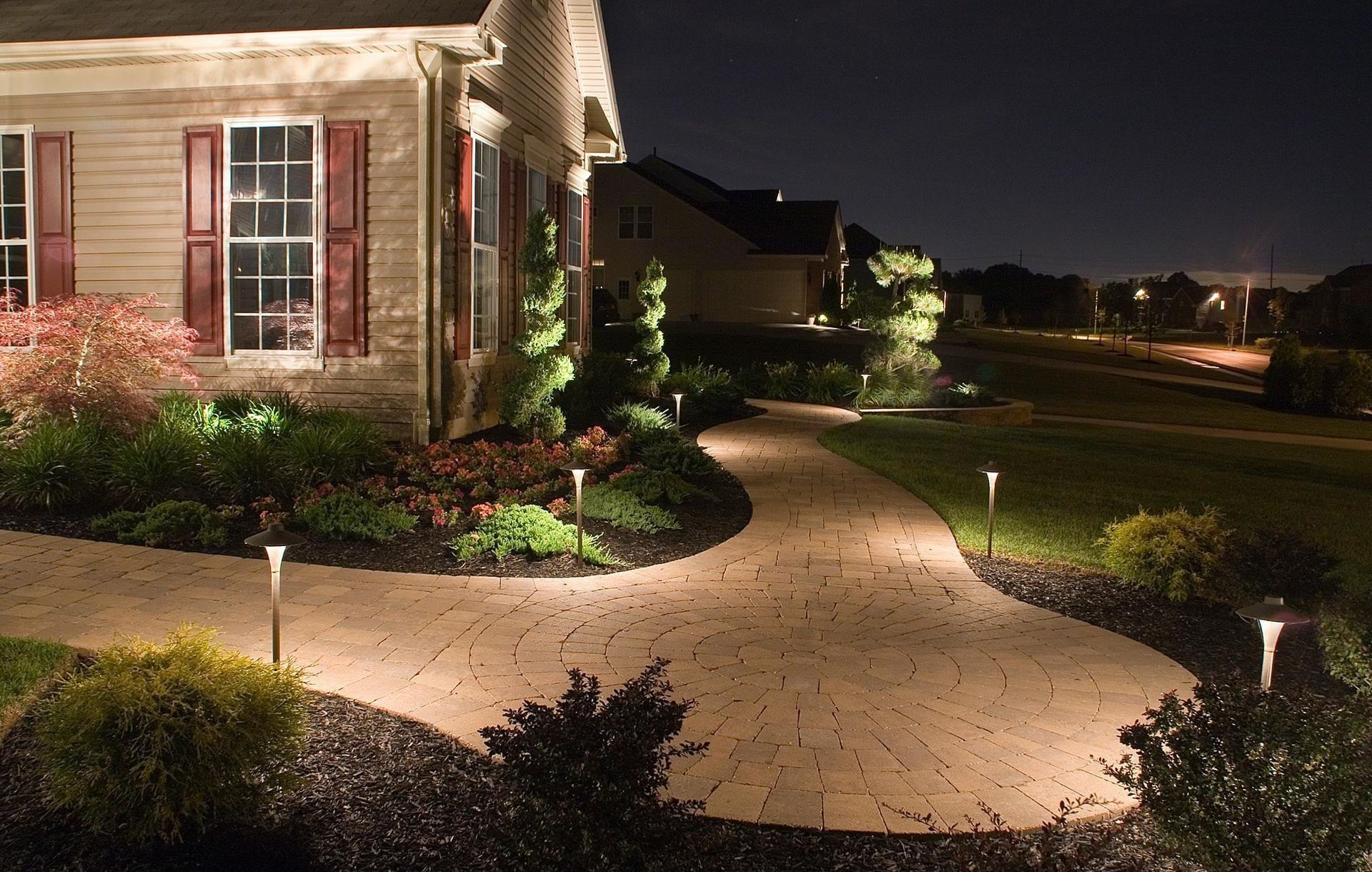 Lit pathway leading to a house at night, surrounded by landscaping and small lights.