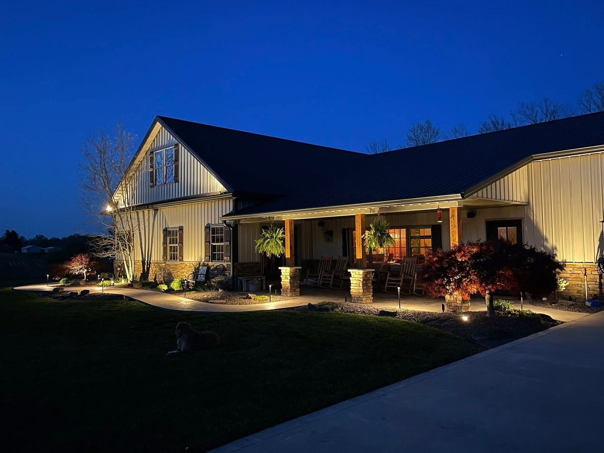 Lit-up, two-story house at night with a porch, landscaping, and a dark blue sky.