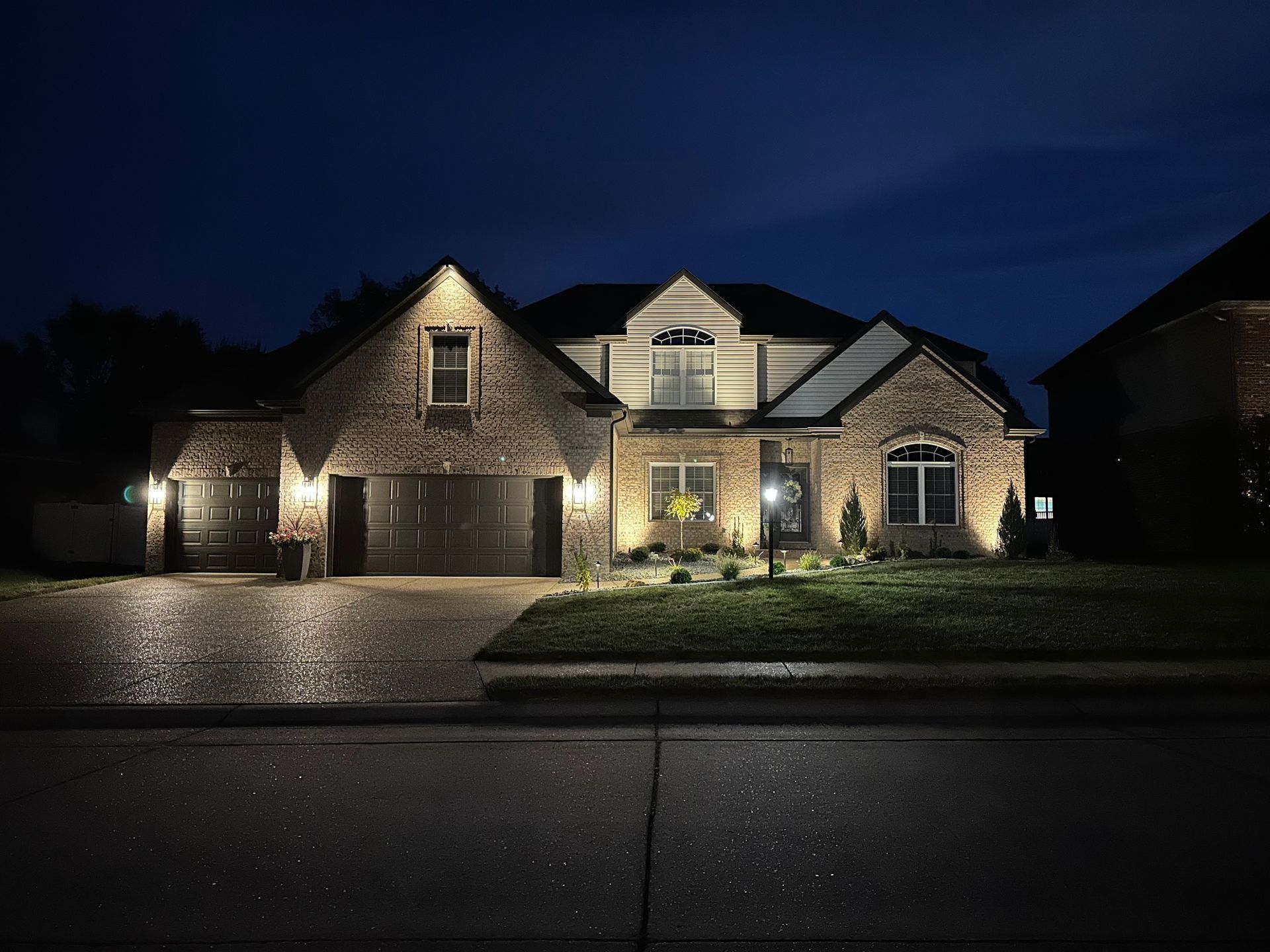 A brick house at night, lit by outdoor lighting.  The driveway and street are wet.