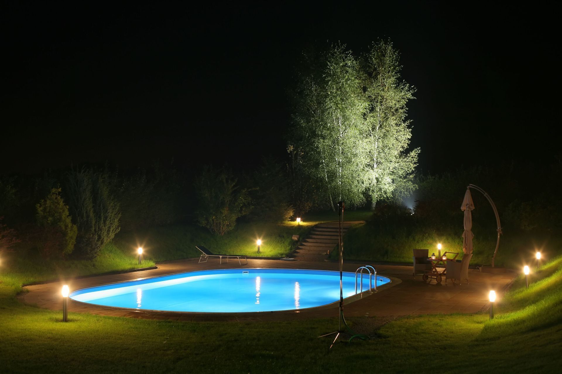 Pool at night, illuminated by lights, with a tree in the background.
