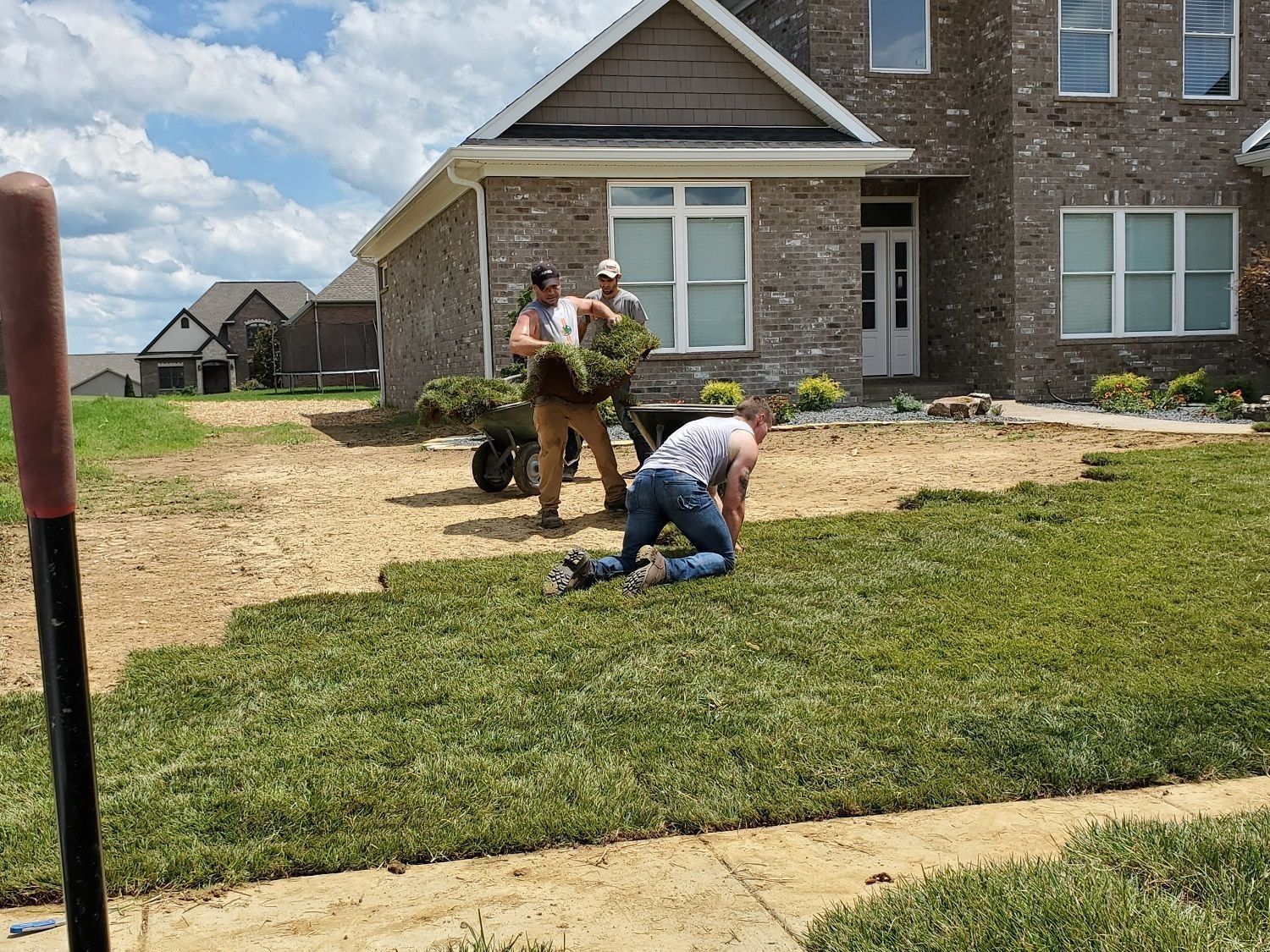 Men laying sod in front of a brick house; one kneels, others carry grass, cloudy sky.