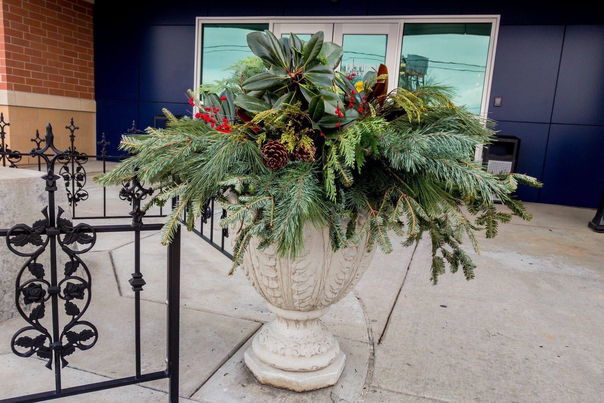 A large stone urn overflowing with winter greenery and red berries sits in front of a building.