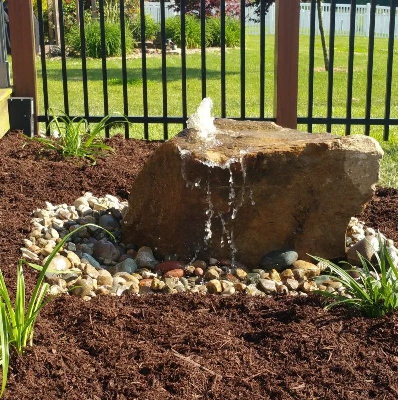 Rock fountain in a garden, water spraying from the top. Surrounded by mulch, pebbles, and green plants, with a fence in the background.