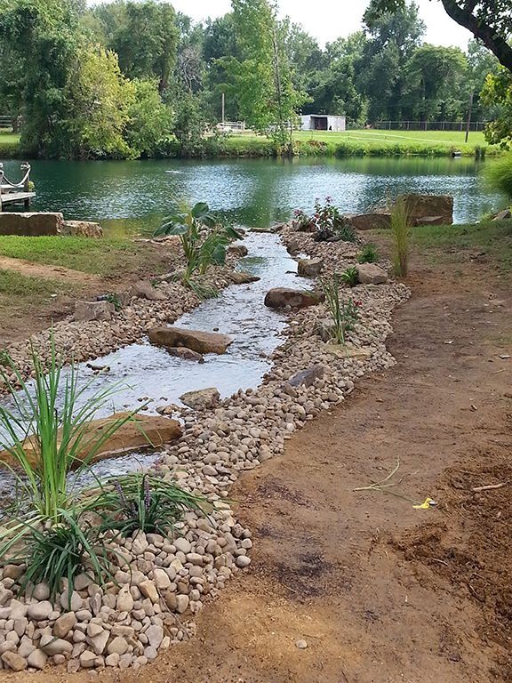 A stream of water flows through a rocky bed towards a calm lake. Plants line the edges, with trees in the background.