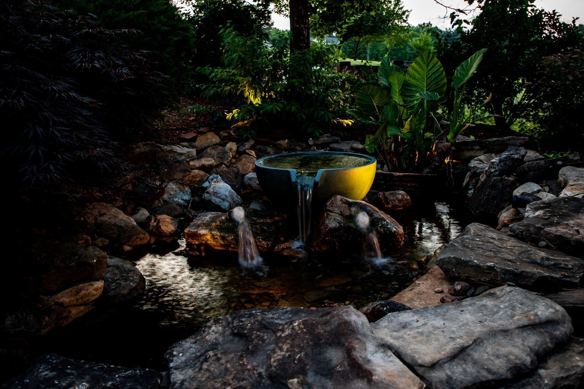 Water fountain with a yellow bowl, surrounded by rocks and lush green plants.