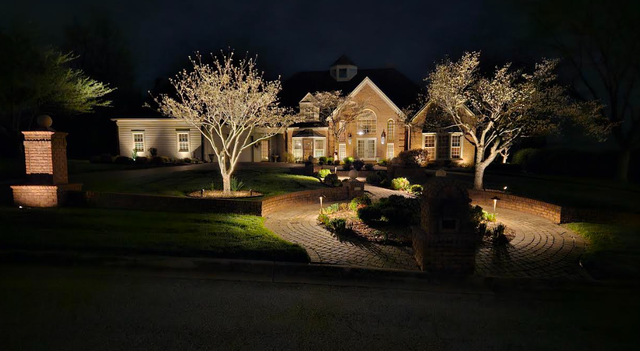 Night view of a brick house with landscape lighting illuminating trees, pathways, and pillars.
