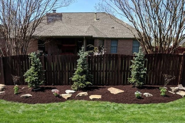 Brown fence with evergreen trees and a mulch bed in front of a house.