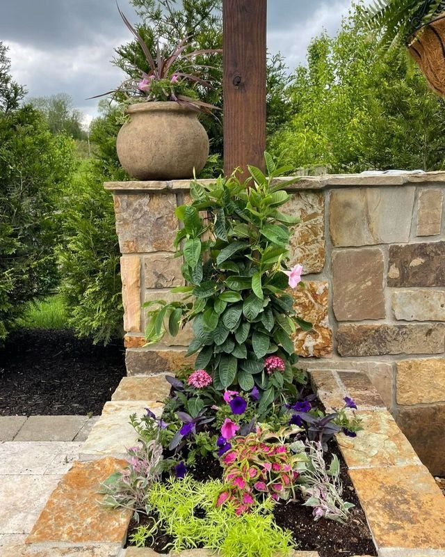 Stone flower bed with colorful flowers and foliage, next to a stone wall. A pot of flowers sits on top.