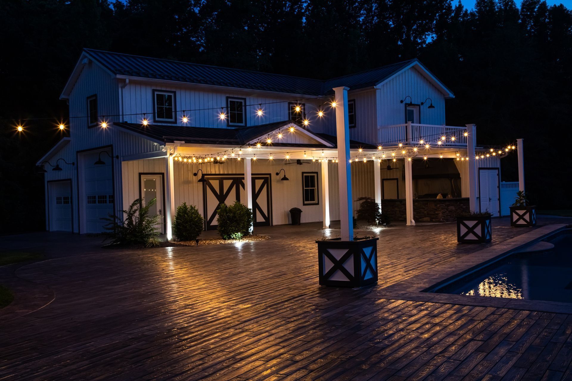 White farmhouse exterior with string lights at dusk, reflecting on brick patio.