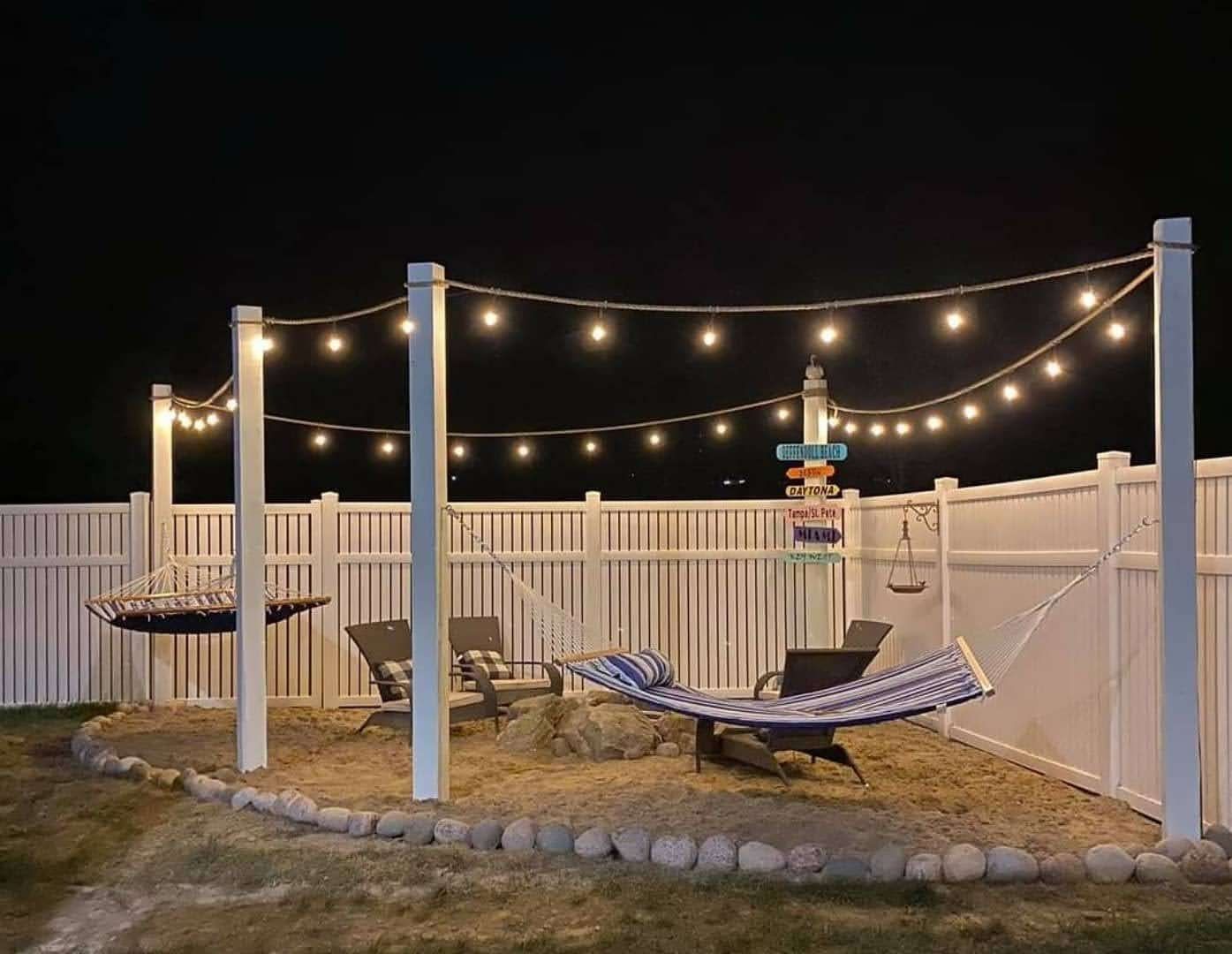 Backyard lounge with string lights, hammocks, chairs, and a fence at night.