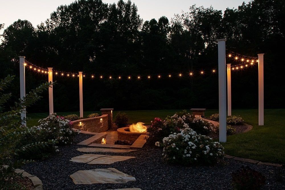 Outdoor fire pit area with string lights, surrounded by landscaping and trees at dusk.