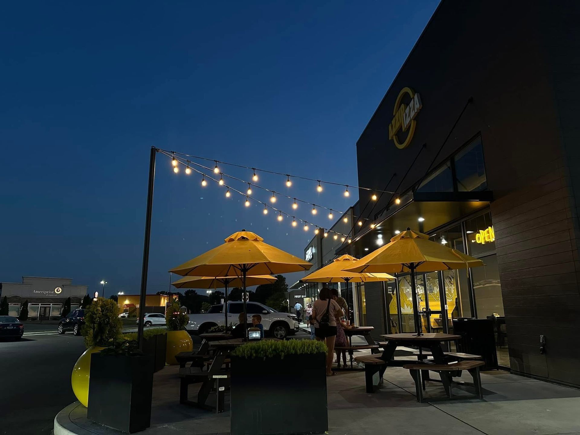 Nighttime outdoor dining area with yellow umbrellas, string lights, and black building.