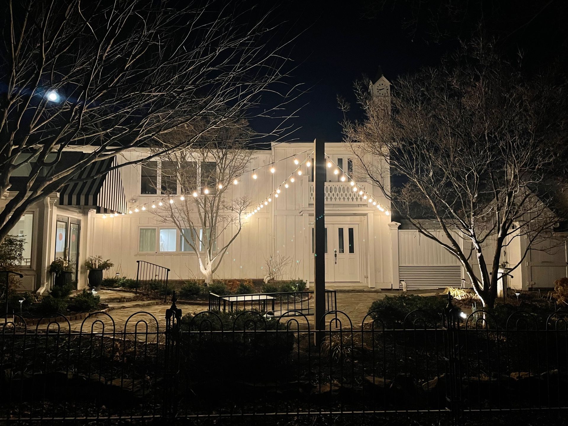A white building at night with string lights, bare trees, and a small fence in front.