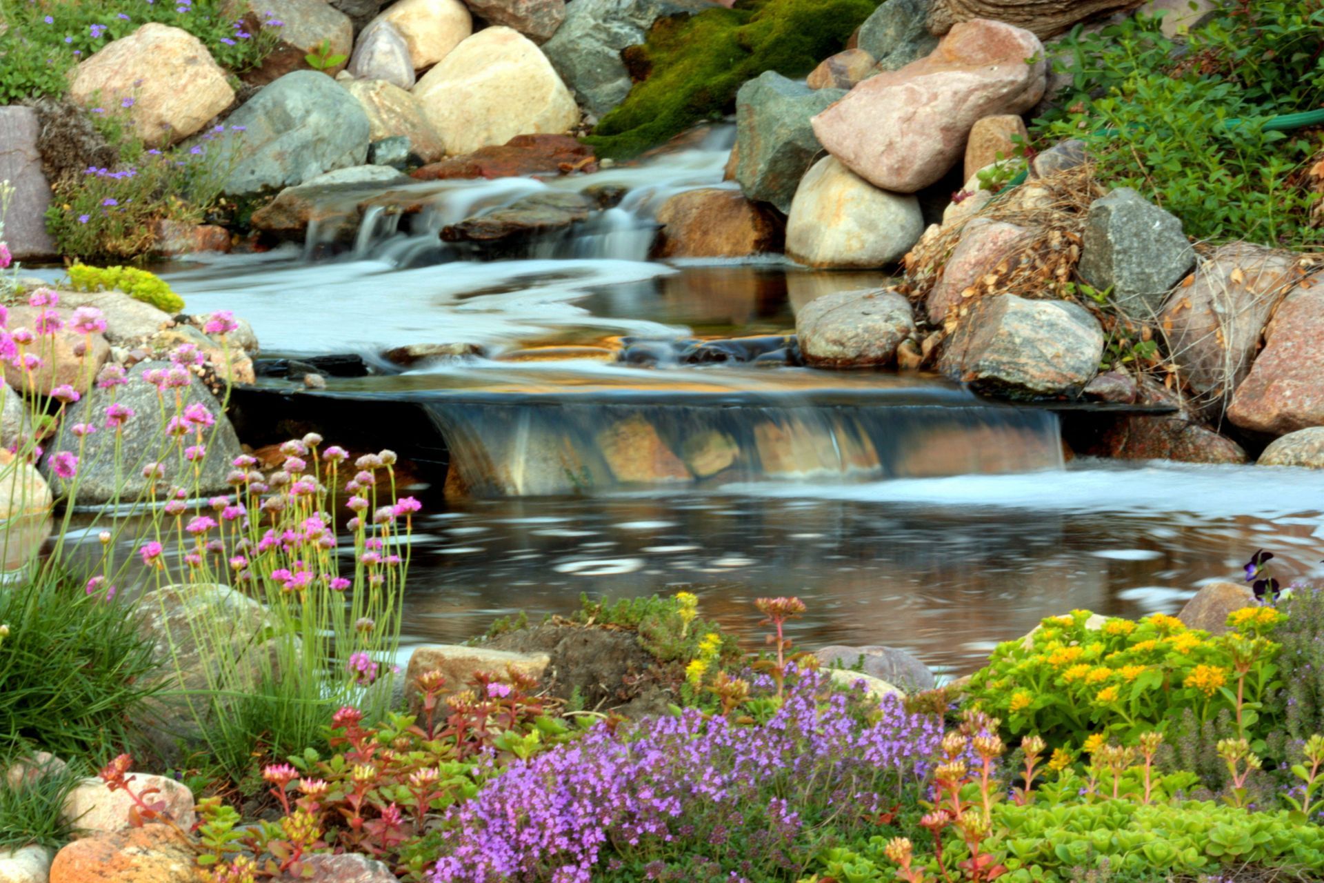A flowing waterfall in a garden, surrounded by rocks and colorful flowers.