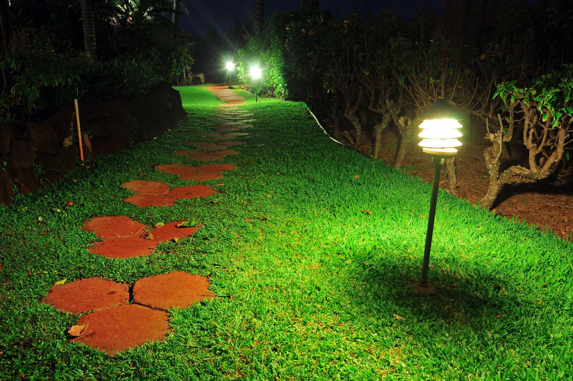 A garden path lit by lamps at night, with stepping stones and lush green grass.