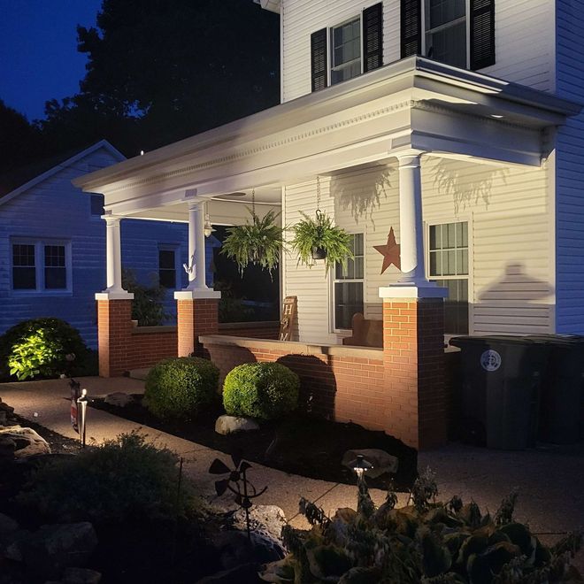 Lit-up white house porch at night with brick columns, potted plants, and pathway lighting.