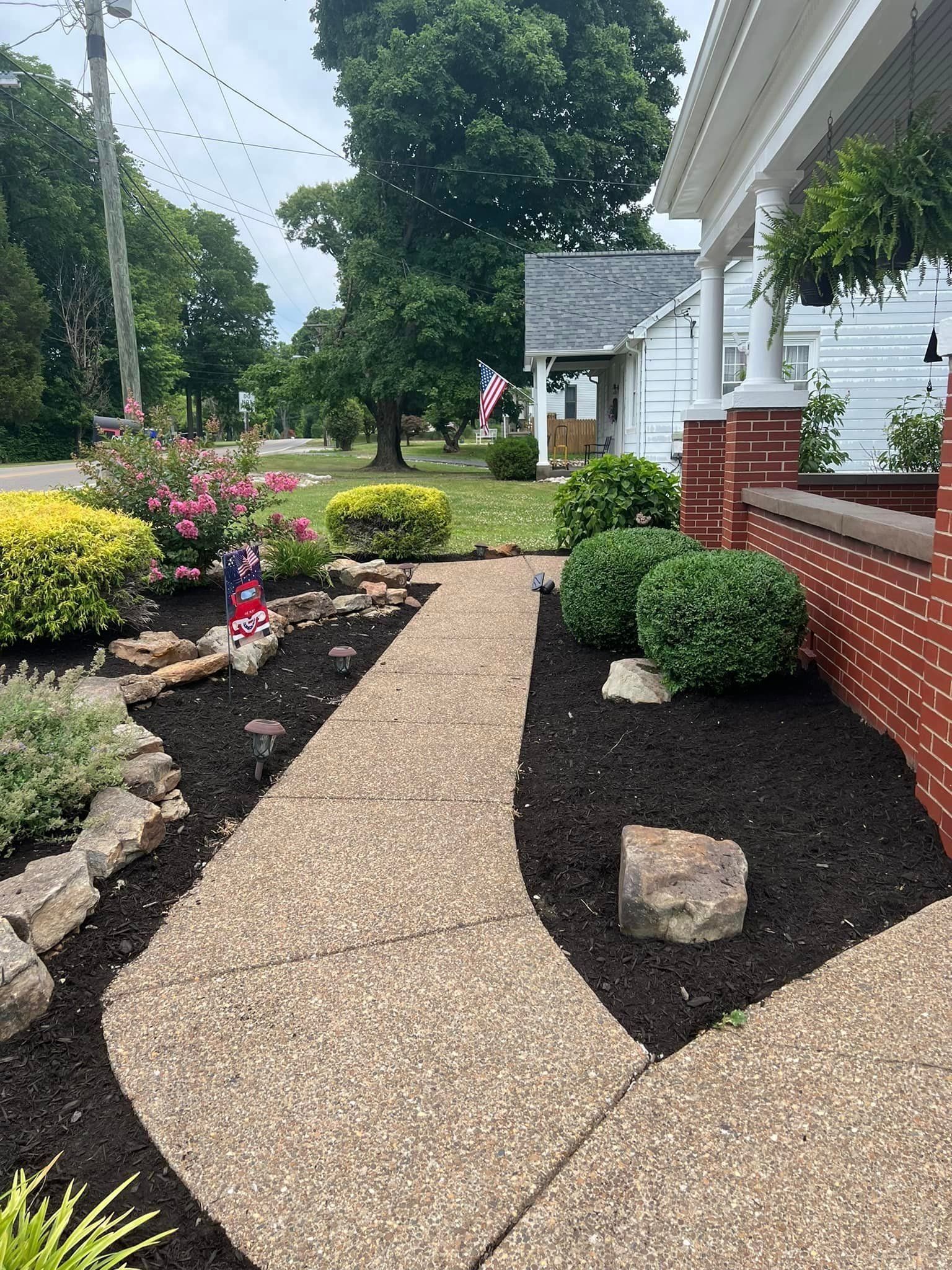 A stone path leads to a white house with hanging plants, surrounded by landscaped flower beds with shrubs.