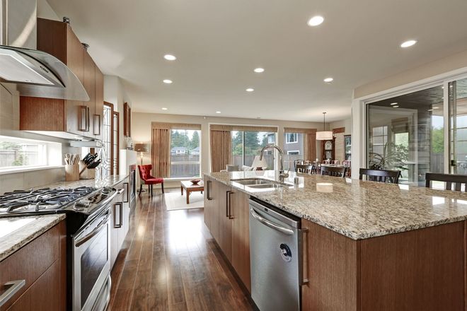 A wide shot of a modern kitchen with wooden cabinets, a granite-topped island, and an open floor plan to a living area.