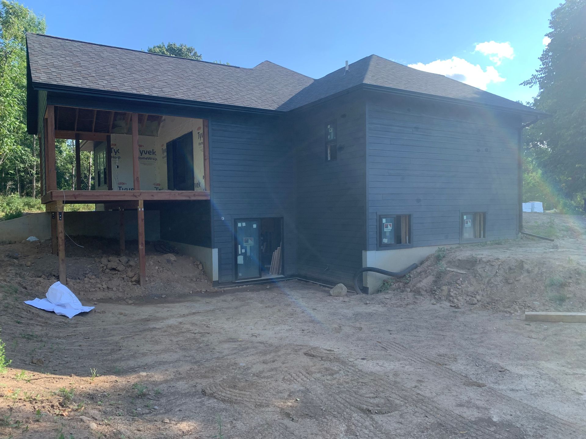 Partially finished dark gray house exterior with porch and dirt yard under a blue sky