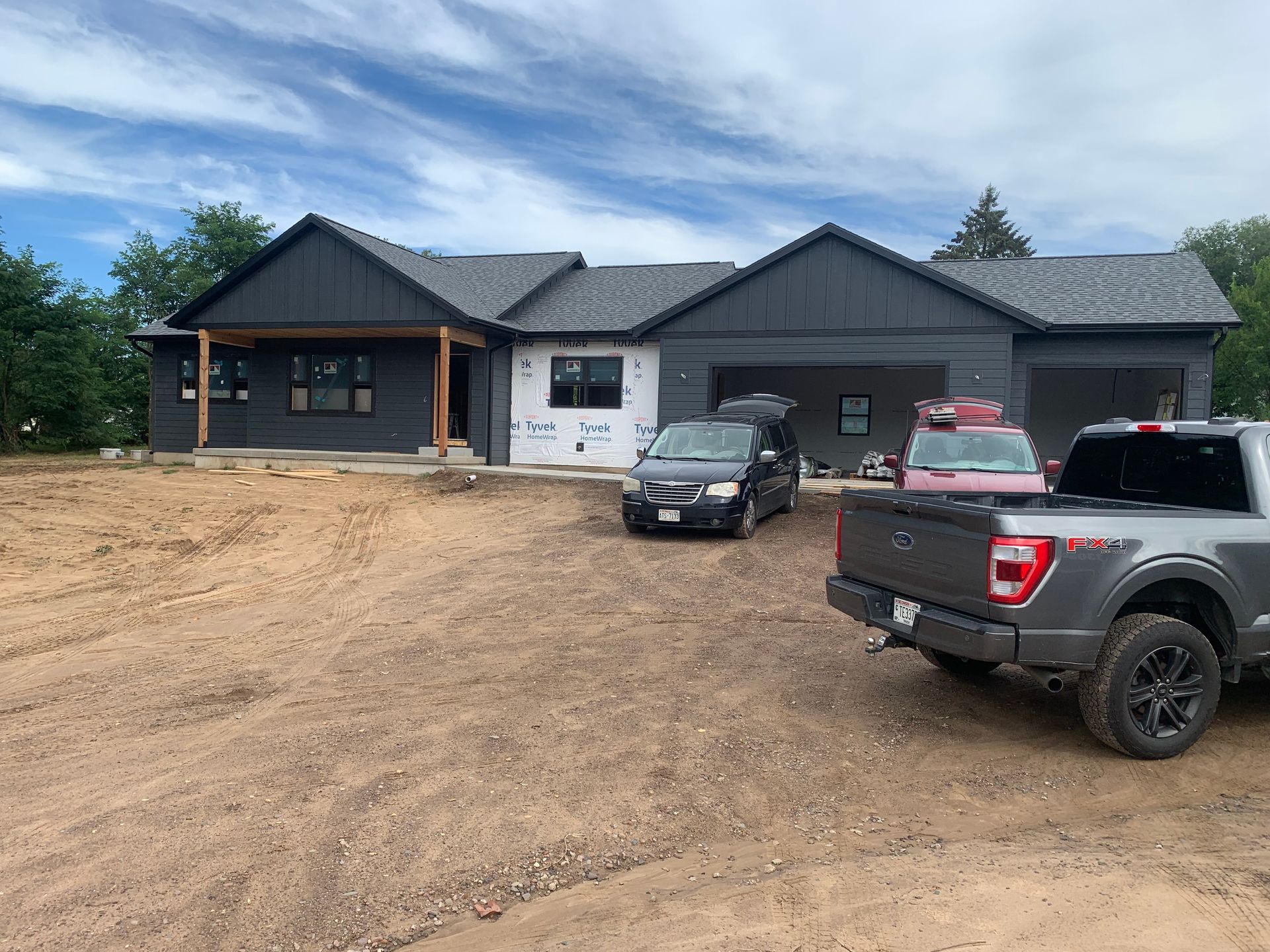 New dark-gray house under construction with trucks parked on a dirt driveway under a blue sky