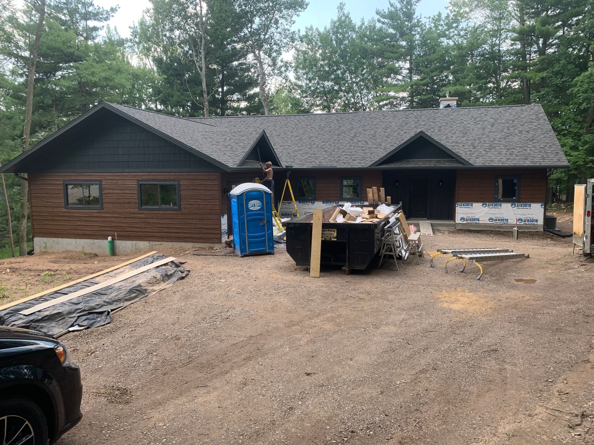 Brown cabin under construction with gravel driveway, blue portable toilet, and materials stacked outside