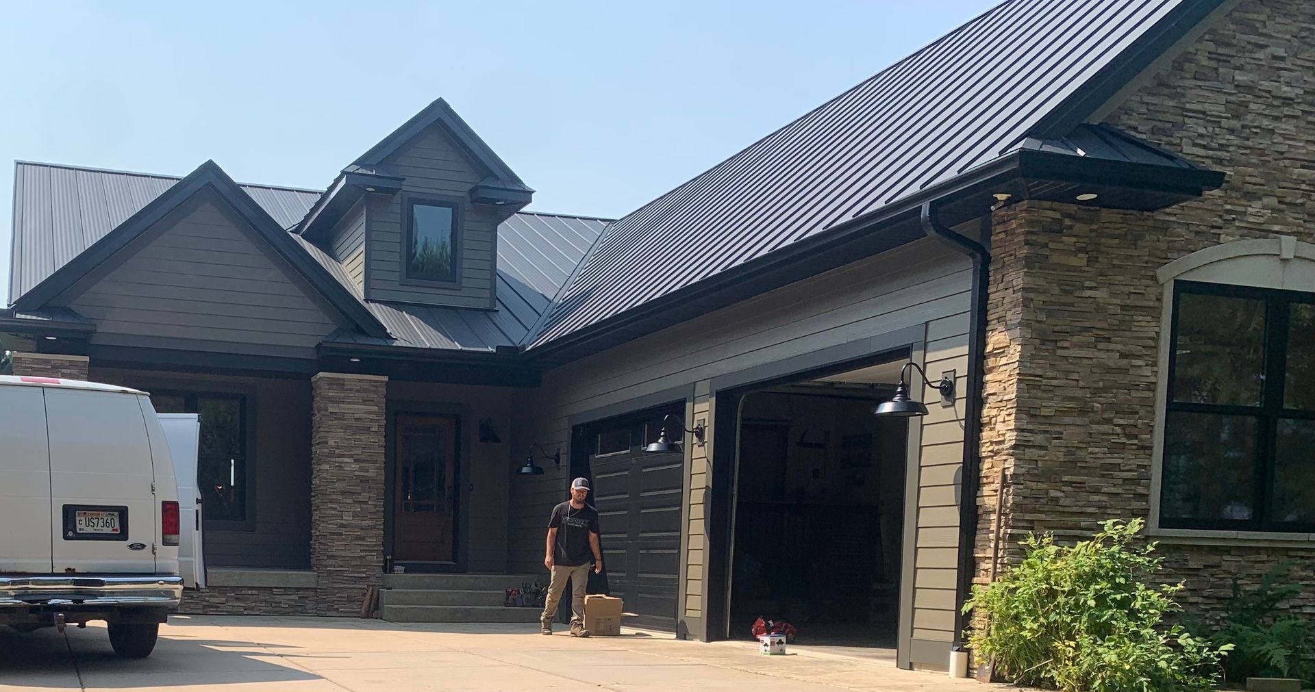 Brick house with a dark roof, open garage, and a person standing in the driveway beside a white van.