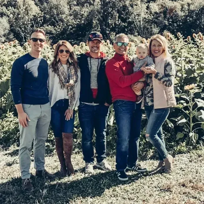 Family standing outdoors in front of tall plants. Several adults and a baby smile at camera.