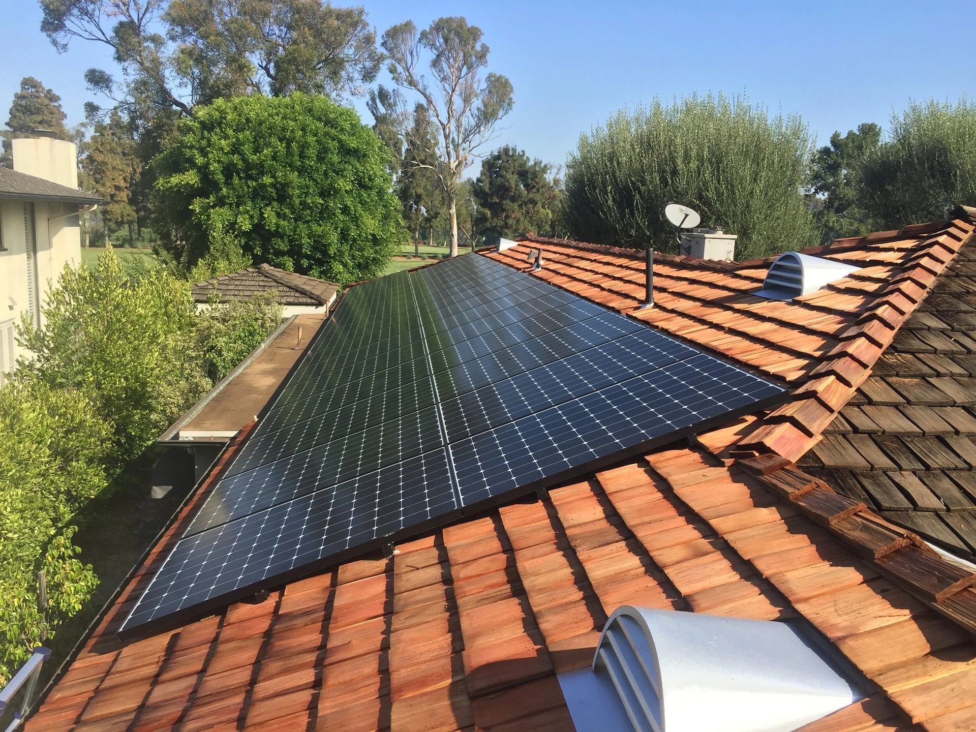 Solar panels installed on a terracotta-tiled roof beside trees and neighboring houses.