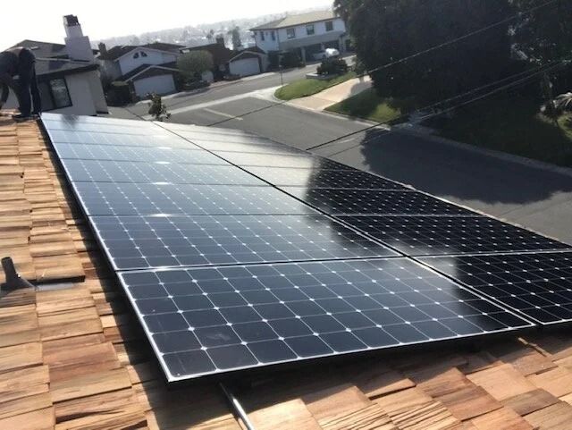 Solar panels mounted on a rooftop beside tan shingles, with a suburban neighborhood in the background.