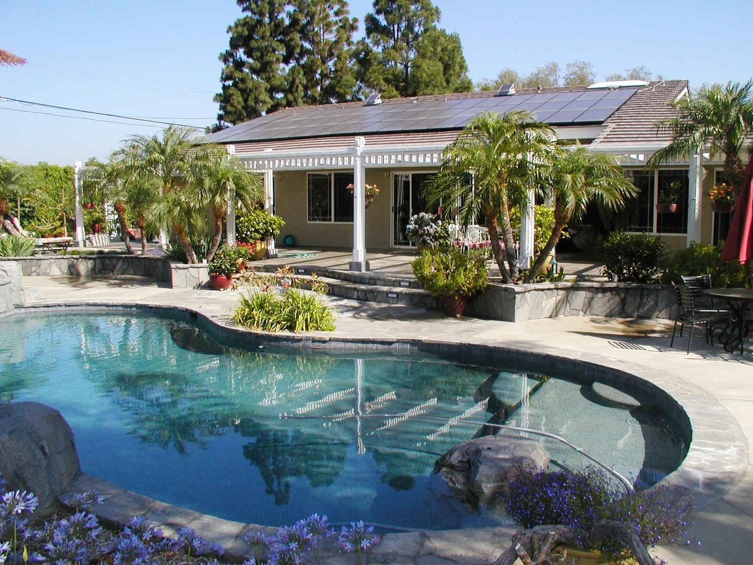 Backyard pool with fountain in front of a house, surrounded by tropical plants and patio seating