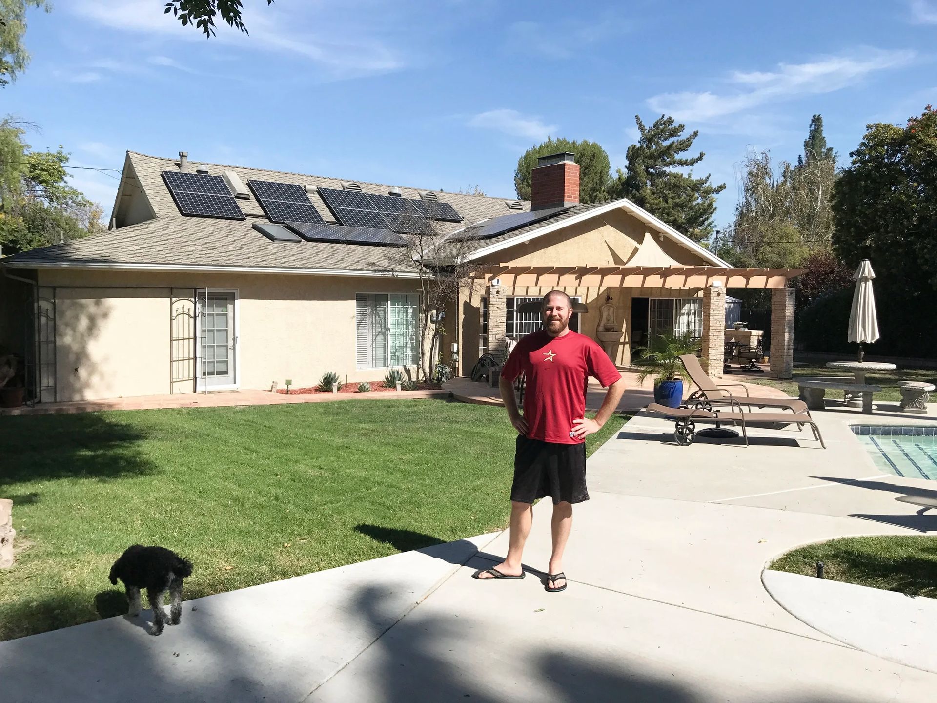 Person standing on a patio beside a pool and house, with a dog in the foreground.
