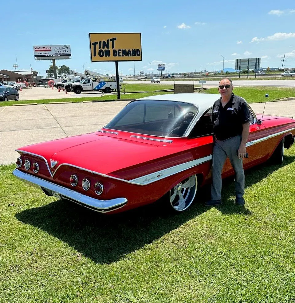 A man is standing next to a red car in front of a yellow sign that says tint on demand.