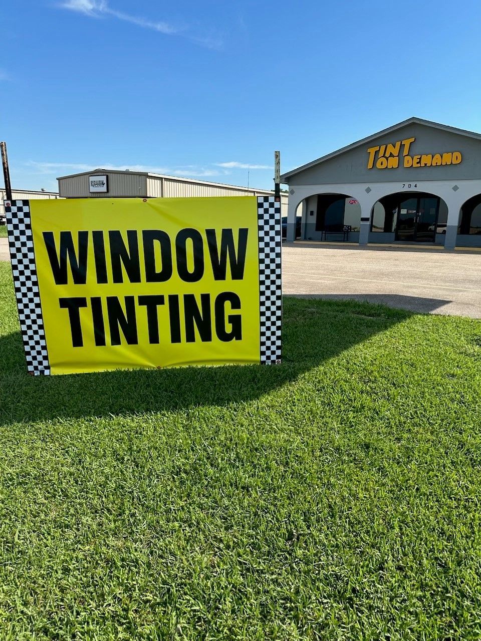 A yellow sign that says window tinting is sitting in the grass in front of a building.