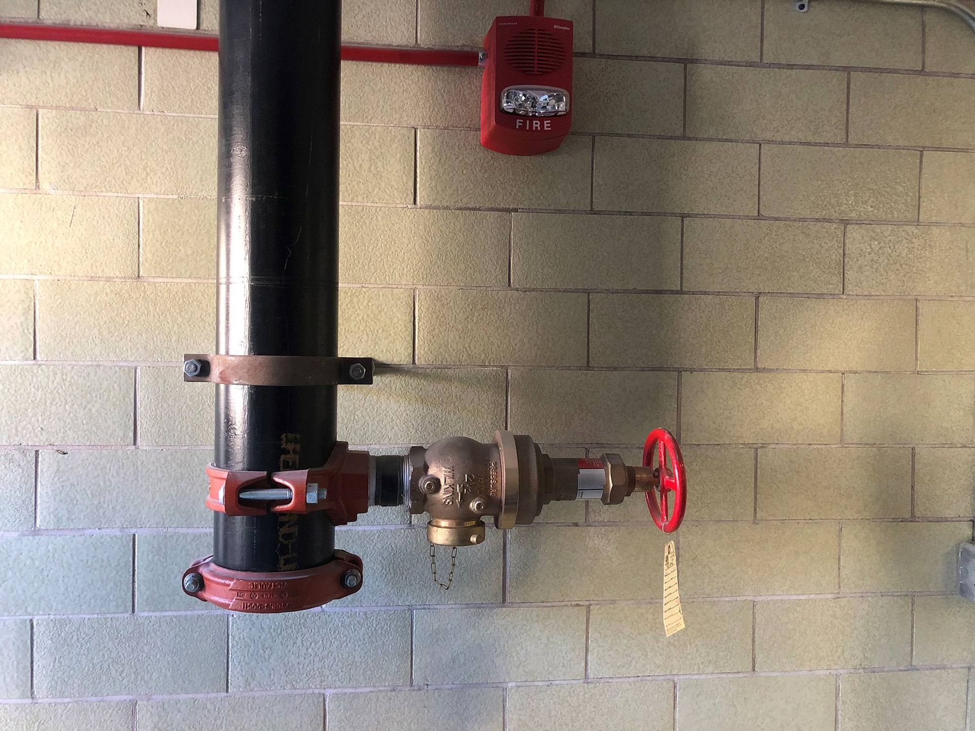 Black pipe and valve attached to a brick wall, with a red fire alarm above.