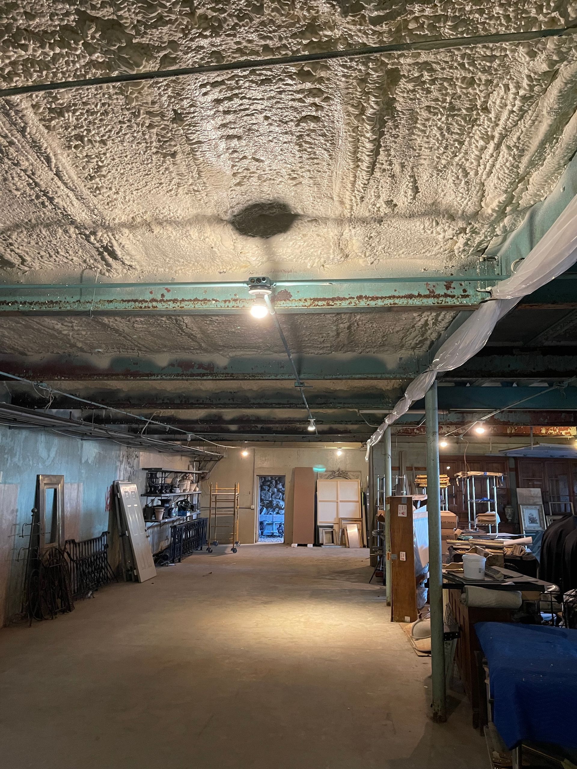 Warehouse interior with exposed ceiling beams and spray foam insulation, containing various furniture and equipment.