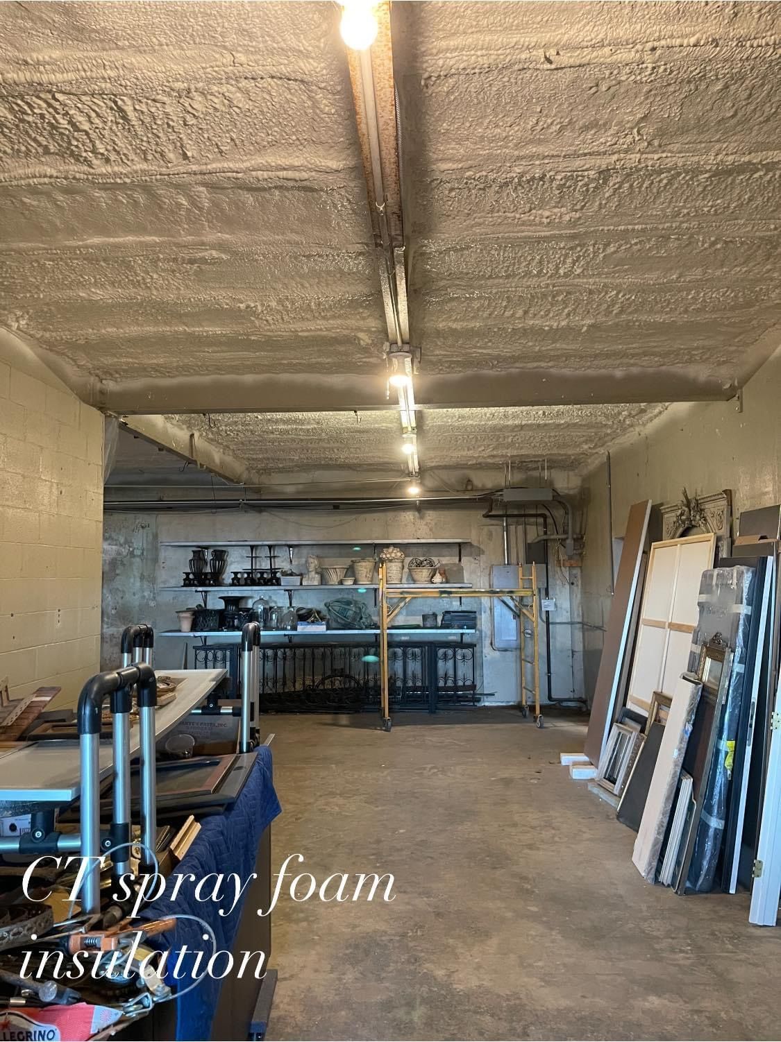 Interior view of a workshop with spray foam insulation on the ceiling, lit by overhead lights. Tools and work tables are visible.