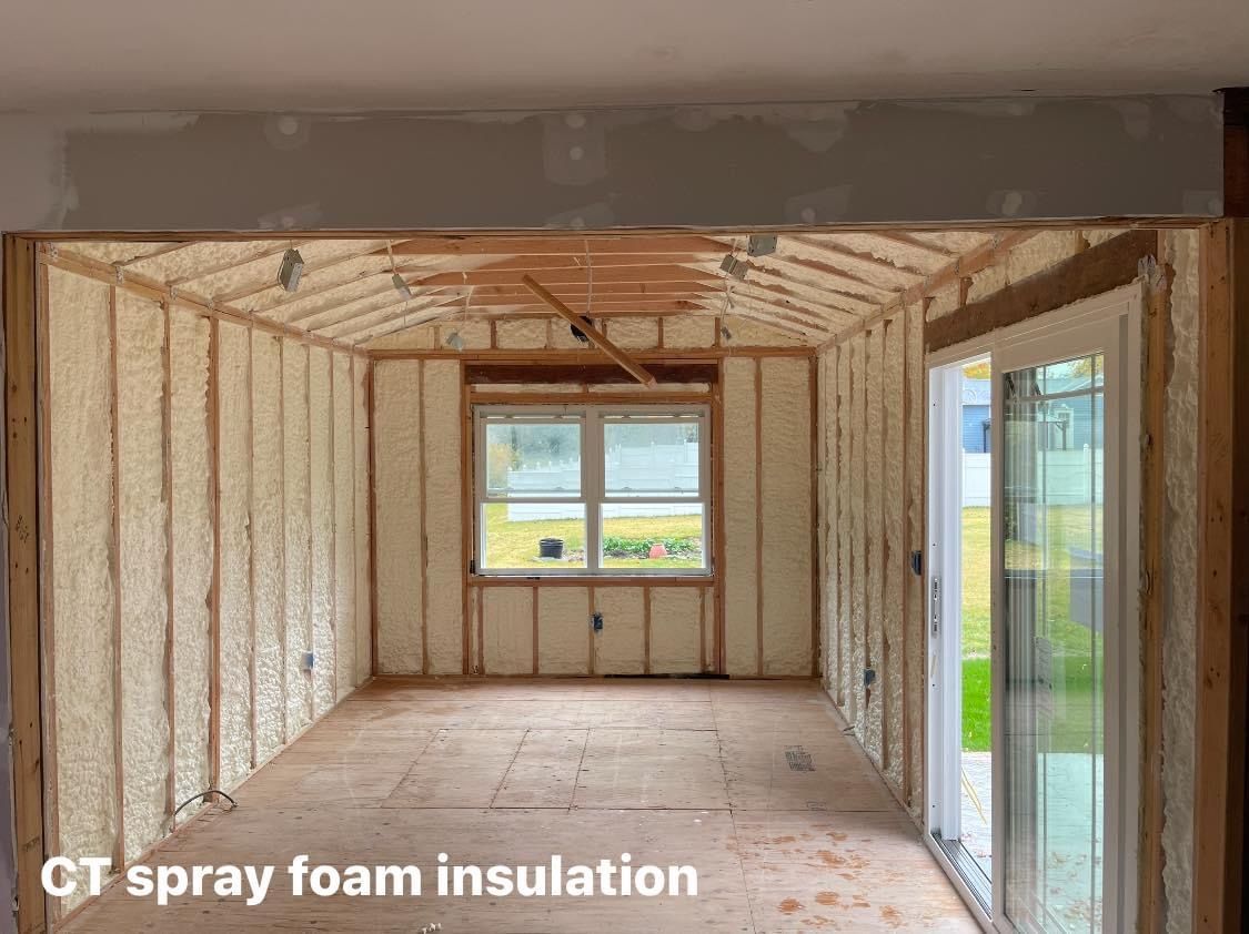 Interior room under construction, walls insulated with light-colored spray foam, window visible, doorway to the right.