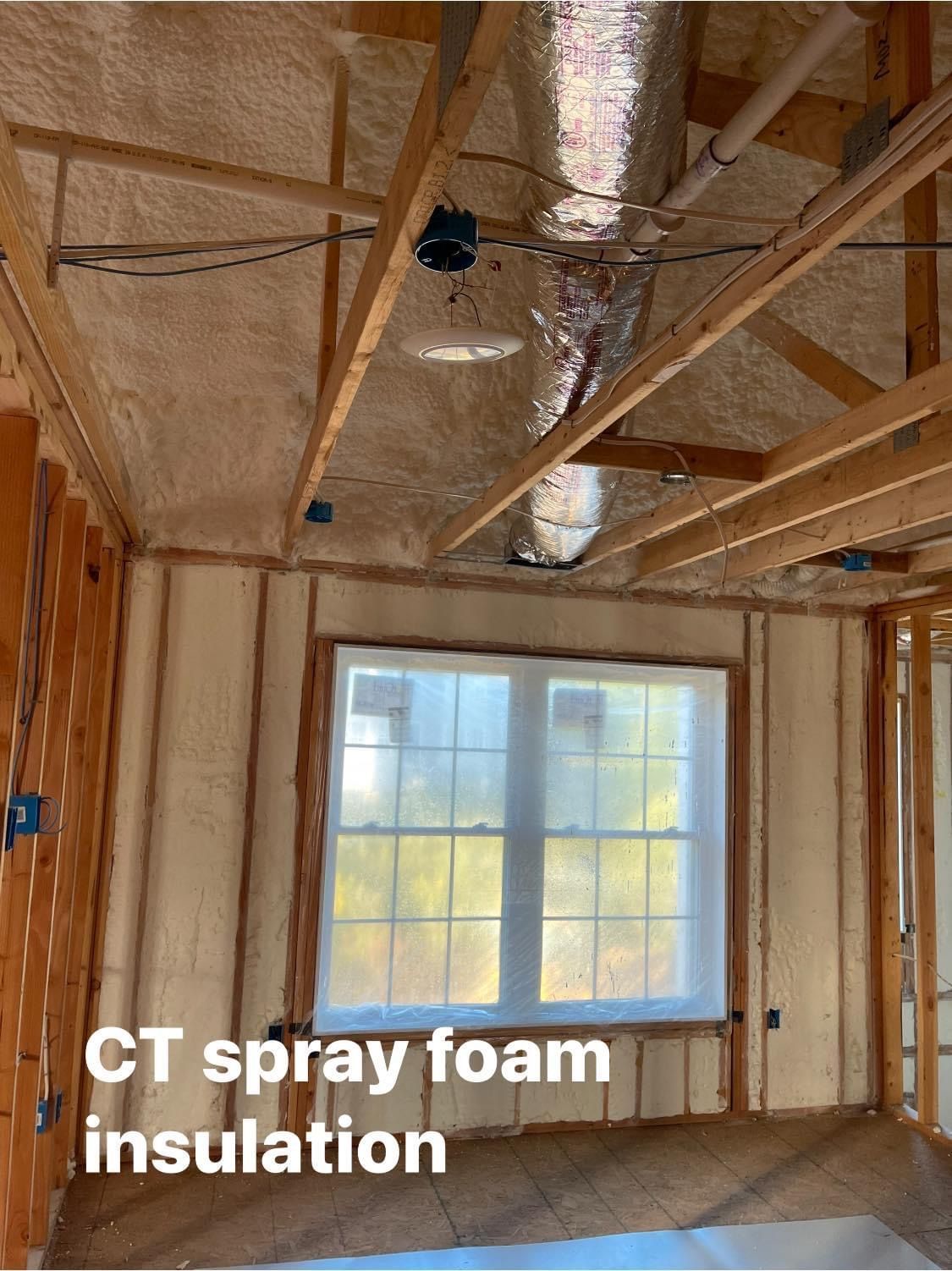 Interior of a room under construction, insulated with spray foam. Window visible, wooden beams and framing.