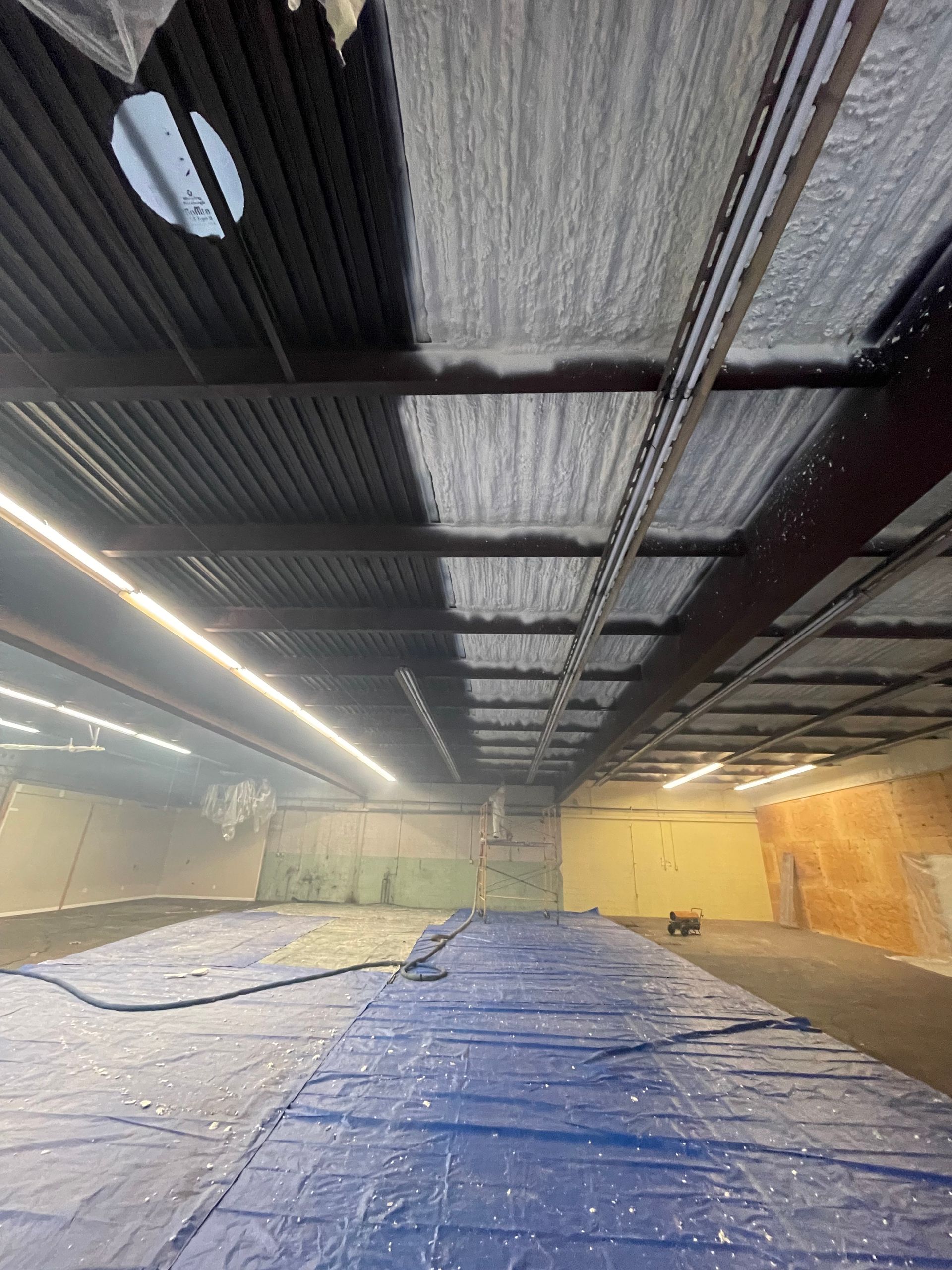 Blue tarp covers a floor, industrial building with dark metal ceiling, covered in sprayed insulation.