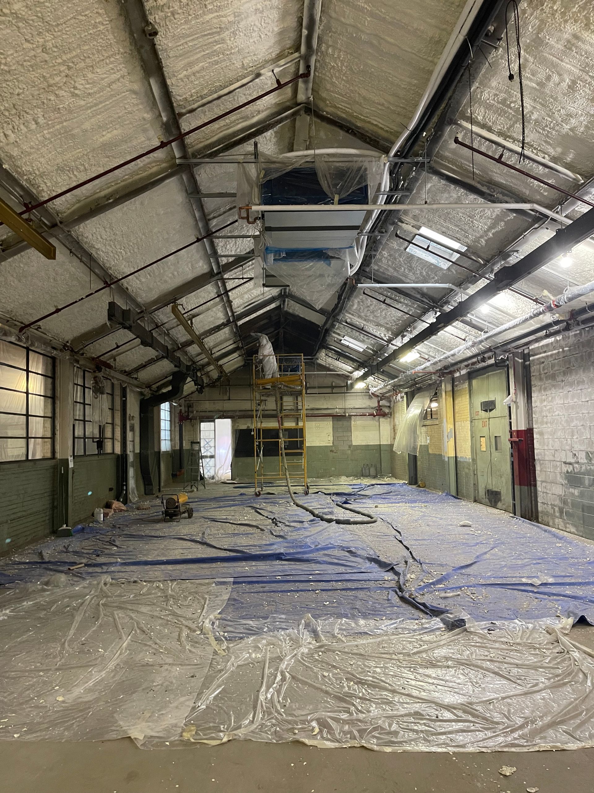 Interior of a warehouse with a metal frame roof, blue tarp on the floor, and insulation on the ceiling.