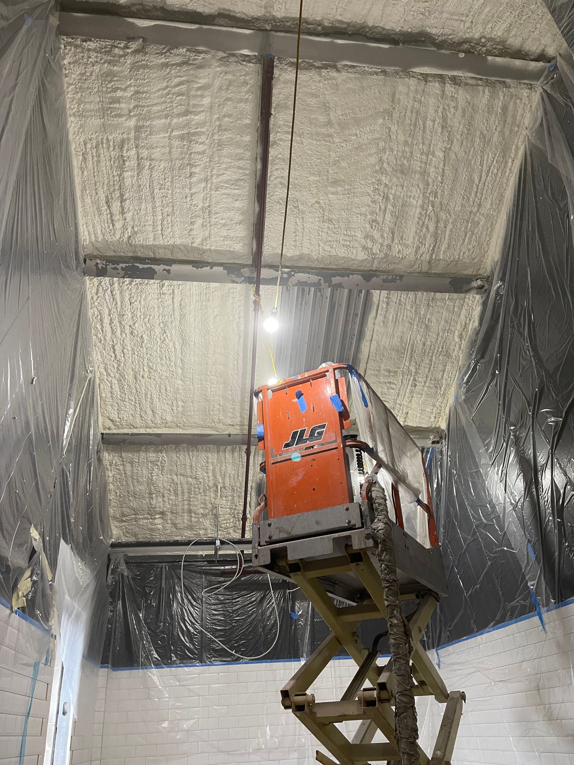 An orange scissor lift reaching a ceiling covered in spray foam insulation. Black plastic sheeting covers the walls.