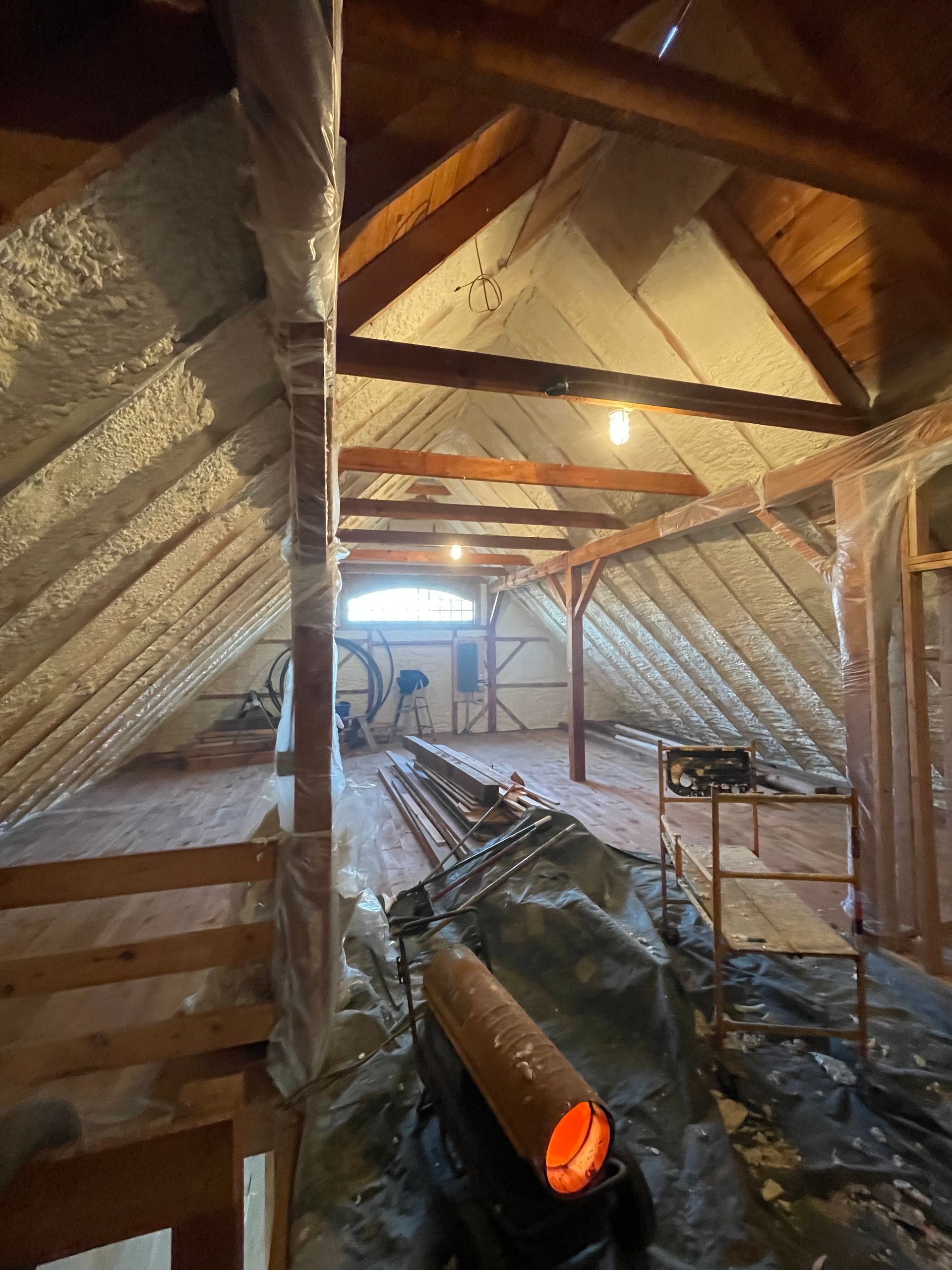 Attic interior with exposed wood beams, spray foam insulation, and construction materials on the floor.