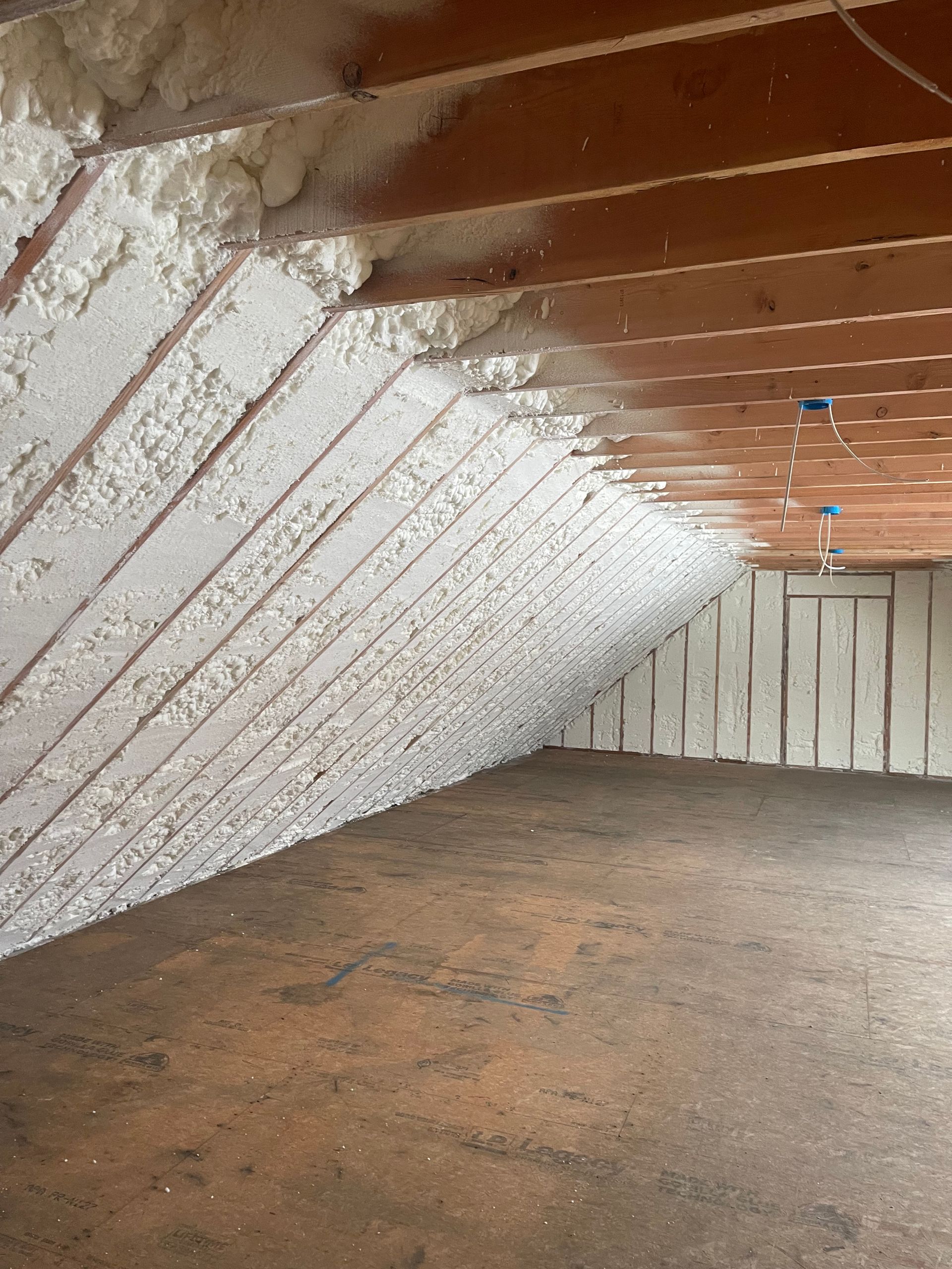 Attic with angled ceiling and walls insulated with white spray foam. Wooden beams and unfinished floor.