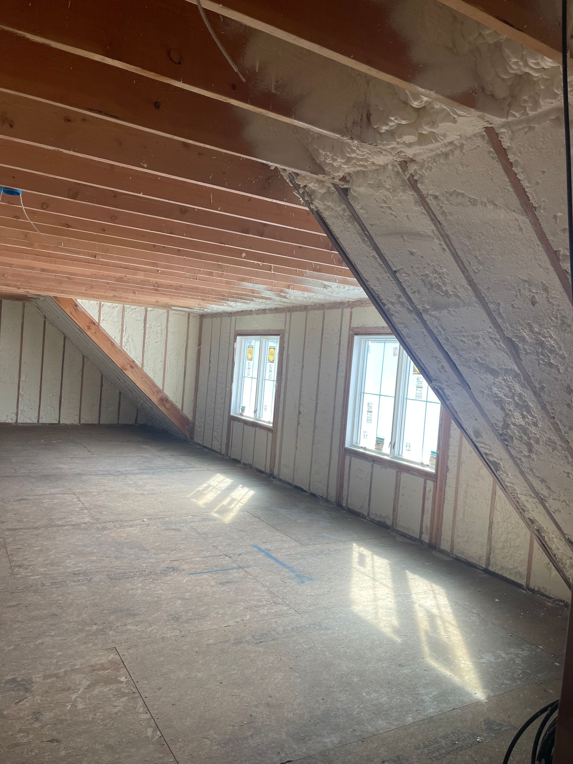 Attic interior with spray foam insulation on walls and rafters. Two windows provide light onto plywood floor.