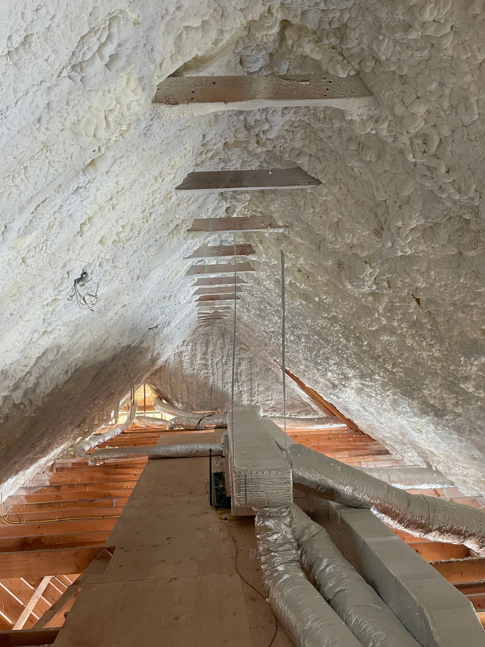 Attic interior with spray foam insulation on the roof and beams. Ducts and wood supports are visible.