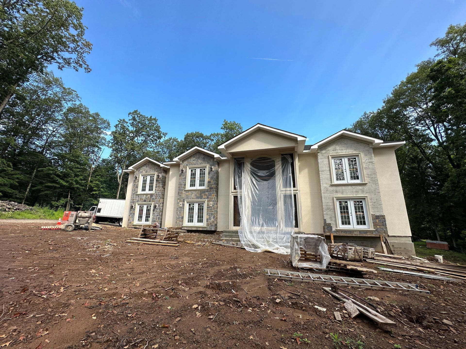 New house under construction; stucco facade, stone accents, clear plastic covering front door, blue sky.