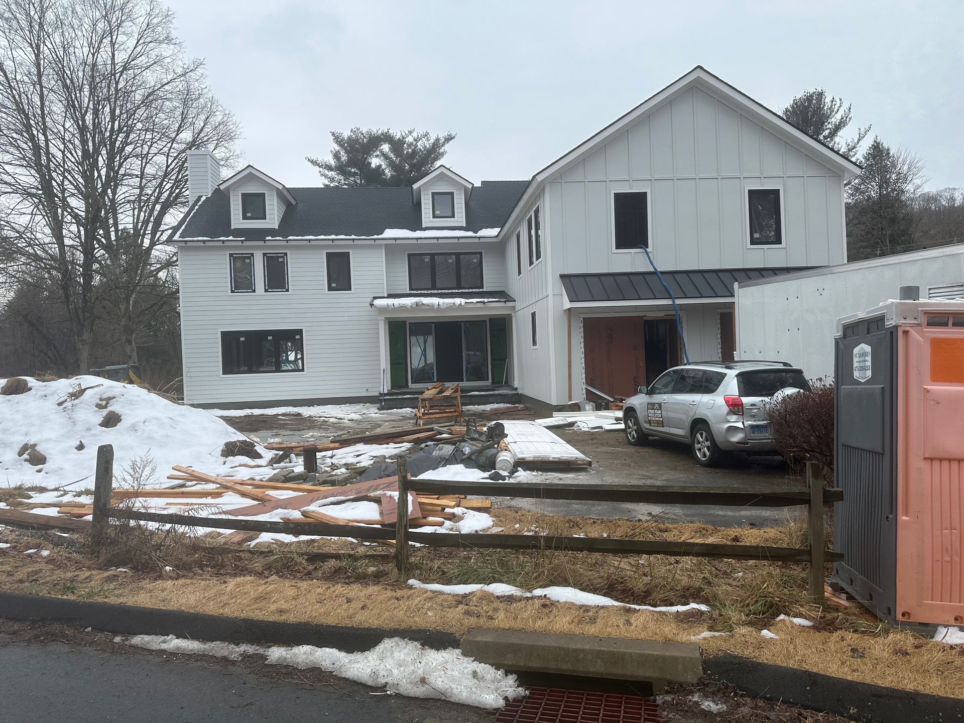 White farmhouse under construction with black roof, windows, and a partially snow-covered yard.