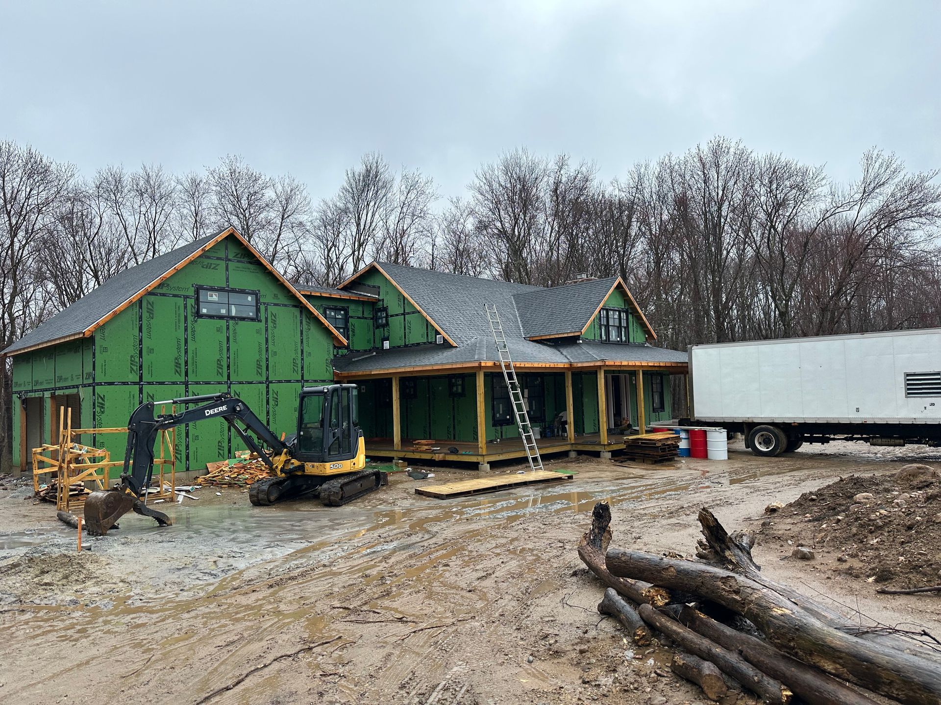 Construction site: partially built house with green siding, excavator, and trailer. Overcast sky.